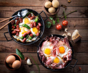 Pan with fried eggs with traditional Spanish iberian ham next to a pan with fried eggs, peppers and chorizo served on rustic wooden table with kitchen utensils and products - ADSF12934