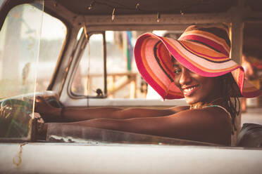 Side view of cheerful young African American female in colorful wide brimmed striped hat sitting behind wheel of vintage van and looking at camera - ADSF12978