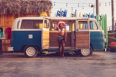 Junge afroamerikanische Besitzerin eines örtlichen Surfbrettverleihs mit buntem Bikinioberteil und Hut neben einem alten Lieferwagen mit einer Reihe von Surfbrettern im Hintergrund - ADSF12981