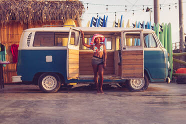 Young African American female owner of local surf board rental wearing colorful bikini top and hat standing next to vintage van with row of surf boards in background - ADSF12981