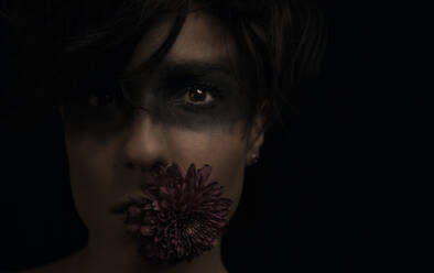 Crop nachdenklich junge dunkelhaarige weibliche Modell mit schwarzer Farbe um die Augen und Blume im Mund Blick auf die Kamera im Studio auf schwarzem Hintergrund - ADSF12983