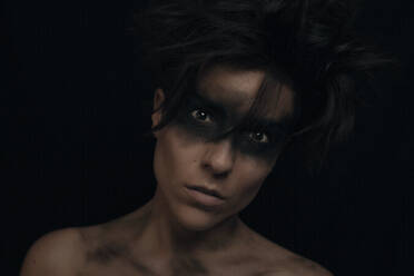 Nachdenkliches junges dunkelhaariges weibliches Modell mit ungekämmtem Haar und schwarzem Make-up um die Augen, das in die Kamera im Studio auf schwarzem Hintergrund schaut - ADSF12984