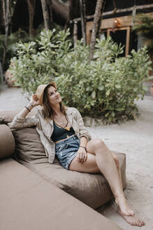 Lächelnde Frau mit Hut sitzt auf einer Couch im Freien - SMSF00177