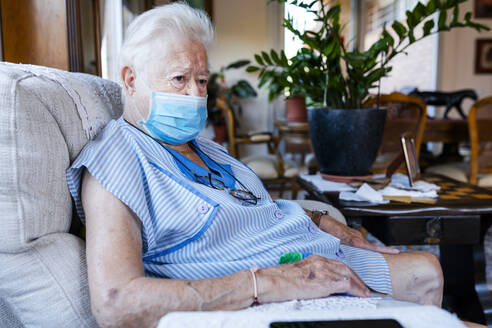 Female senior with protective mask sitting in armchair at home - GEMF04094
