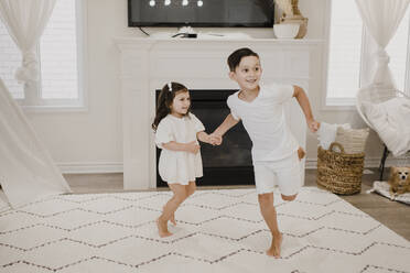 Verspielte Geschwister, die sich beim Laufen im Wohnzimmer an den Händen halten - SMSF00200