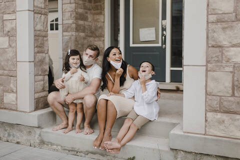 Smiling family with kids wearing protective face mask while sitting on steps outside house stock photo