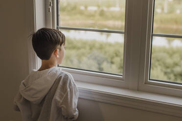 Junge schaut durch das Fenster, während er zu Hause steht - SMSF00230
