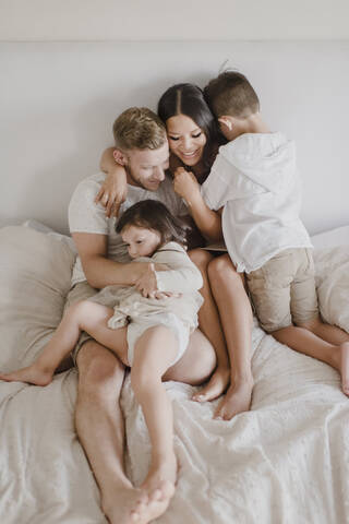 Smiling parents embracing kids while sitting on bed in bedroom stock photo