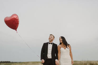 Happy bride and groom with heart shape balloon against sky - SMSF00288