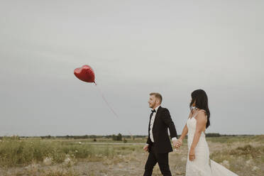 Bride and groom with heart shape balloon holding hands while walking against clear sky - SMSF00289