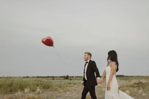 Braut und Bräutigam mit Herzform Ballon halten die Hände beim Gehen gegen den klaren Himmel - SMSF00289