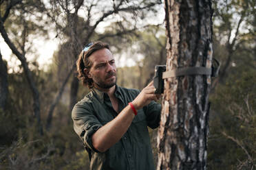 Man adjusting the settings in trail camera mounting on tree trunk in forest - SASF00005