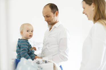 Father dressing up baby boy standing by mother at home - SAJF00087