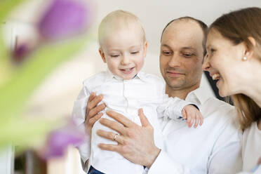 Lächelnder Vater, der einen kleinen Jungen hält, während er neben einer fröhlichen Mutter zu Hause steht - SAJF00092