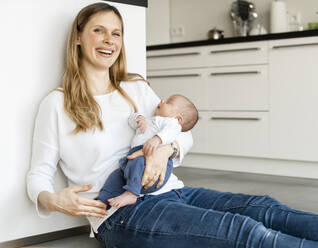Cheerful mother holding sleeping baby boy while sitting on kitchen floor at home - SAJF00103