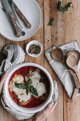 Top view of anonymous person putting hot bowl full of delicious chicken with Parmesan and basil leaves roasted in tomato sauce on lumber table near spices and utensils - ADSF13027