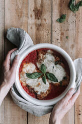 Top view of anonymous person putting hot bowl full of delicious chicken with Parmesan and basil leaves roasted in tomato sauce on lumber table near spices and utensils - ADSF13028