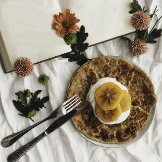 Top view of homemade crepes with cream and sliced persimmon served on plate with knife and fork on surface decorated with white cloth and flowers next to open book with empty pages - ADSF13041