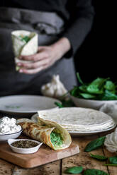 From above of tasty tortilla with puree of fresh spinach on wooden cutting board with faceless lady cooking on blurred background - ADSF13046