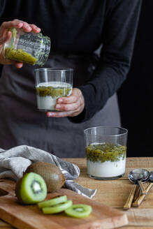 Anonyme Hausfrau in Schürze gibt frische pürierte Kiwi in ein Glas mit Joghurt, während sie ein gesundes Frühstück am Holztisch in der Küche zubereitet - ADSF13059