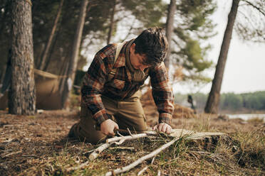 Bushcrafter cutting stick while kneeling in forest - SASF00017