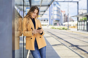 Lächelnde Frau, die ein Smartphone benutzt, während sie an der Straßenbahnhaltestelle wartet - BSZF01610