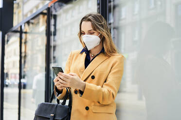 Woman in protective face mask using smart phone while waiting for public transportation - BSZF01616