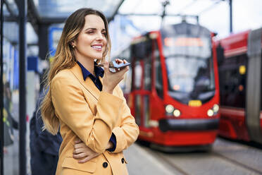 Lächelnde Frau, die einen Lautsprecher benutzt, während sie über ein Mobiltelefon an einer Straßenbahnhaltestelle spricht - BSZF01617