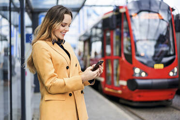 Frau schickt SMS über ihr Smartphone, während sie an der Straßenbahnhaltestelle wartet - BSZF01618