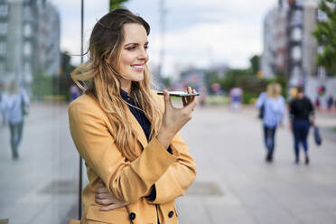 Lächelnde Frau, die an einer Straßenbahnhaltestelle einen Lautsprecher benutzt und über ihr Smartphone spricht - BSZF01620