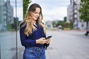 Woman text messaging through mobile phone while standing on footpath in city - BSZF01623