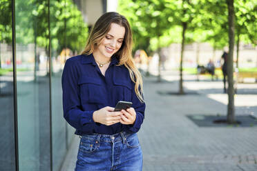 Lächelnde schöne Frau Text-Messaging durch Smartphone beim Stehen auf Gehweg in der Stadt - BSZF01625