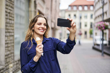 Smiling beautiful woman taking selfie while eating ice cream on footpath - BSZF01628