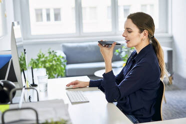 Kreative Geschäftsfrau, die einen Lautsprecher benutzt, während sie über ihr Smartphone im Büro spricht - BSZF01632