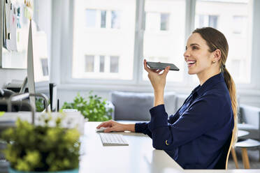 Smiling businesswoman using speaker while talking on smart phone at desk in office - BSZF01633