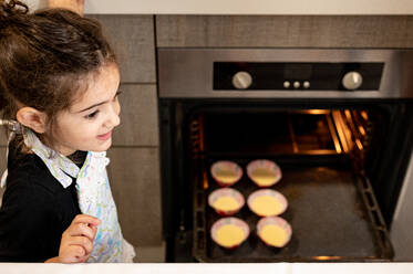 From above adorable ethnic girl in apron standing near open oven with raw cupcakes and looking away while cooking pastry in modern kitchen at home - ADSF13068