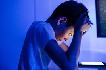 Side view of confused boy watching cellphone empty screen and touching head while sitting in room with blue light at home - ADSF13073