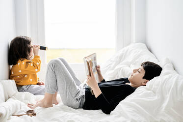 Seitenansicht des Bruders, der ein Buch liest, und der Schwester, die mit einem Retro-Späher spielt und sich auf einem bequemen Bett entspannt und das Wochenende genießt - ADSF13077