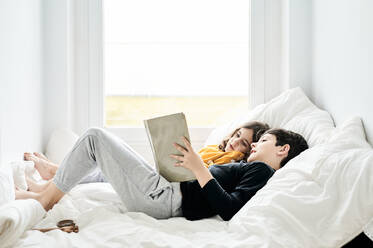 Seitenansicht eines entspannten Geschwisterpaares, das auf einem weichen Bett liegt und gemeinsam ein Märchen liest, während es das Wochenende zu Hause genießt - ADSF13078
