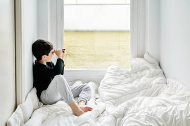 Side view of calm child wearing casual clothing sitting on cozy white blanket in hall and playing with vintage spyglass while looking out of window - ADSF13080