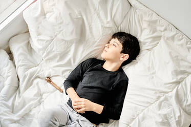 Draufsicht auf ein entspanntes Kind in Freizeitkleidung, das sich auf einer gemütlichen Decke im Schlafzimmer ausruht und aus dem Fenster schaut, während es das Wochenende genießt - ADSF13081