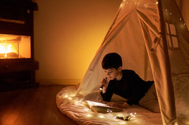 Focused kleines Kind in Freizeitkleidung Lesung Buch beim Liegen in Kinder spielen Zelt mit Taschenlampe und Girlande um in der Nähe von Kamin in Wohnung - ADSF13086