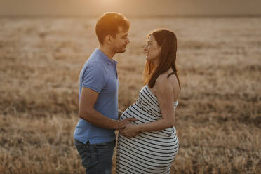 Seitenansicht eines Mannes, der sanft den Bauch einer schwangeren Frau berührt, während sie sich in einem trockenen Feld bei Sonnenuntergang auf dem Lande ansehen - ADSF13088