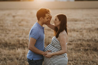 Seitenansicht eines Mannes, der sanft den Bauch einer schwangeren Frau berührt, während sie sich in einem trockenen Feld bei Sonnenuntergang auf dem Lande ansehen - ADSF13089