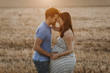 Seitenansicht eines Mannes, der sanft den Bauch einer schwangeren Frau berührt, während sie sich in einem trockenen Feld bei Sonnenuntergang auf dem Lande ansehen - ADSF13090