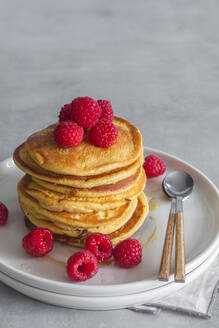 Stapel von leckeren Pfannkuchen mit reifen Himbeeren auf dem Teller in der Nähe von Löffeln auf grauem Hintergrund platziert - ADSF13097