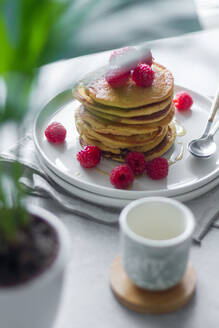 Von oben Teller mit leckeren Pfannkuchen mit Himbeeren auf Serviette neben leerer Tasse und Topfpflanze am Morgen - ADSF13099