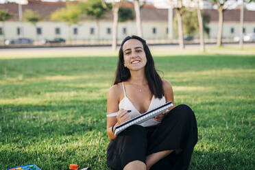 Smiling woman painting while sitting on grass in public park - DCRF00753