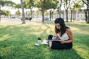 Junge Frau malt Aquarelle in einem Buch, während sie im Park sitzt - DCRF00754