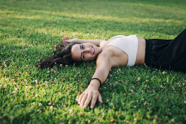 Smiling young woman relaxing on grass at public park - DCRF00759